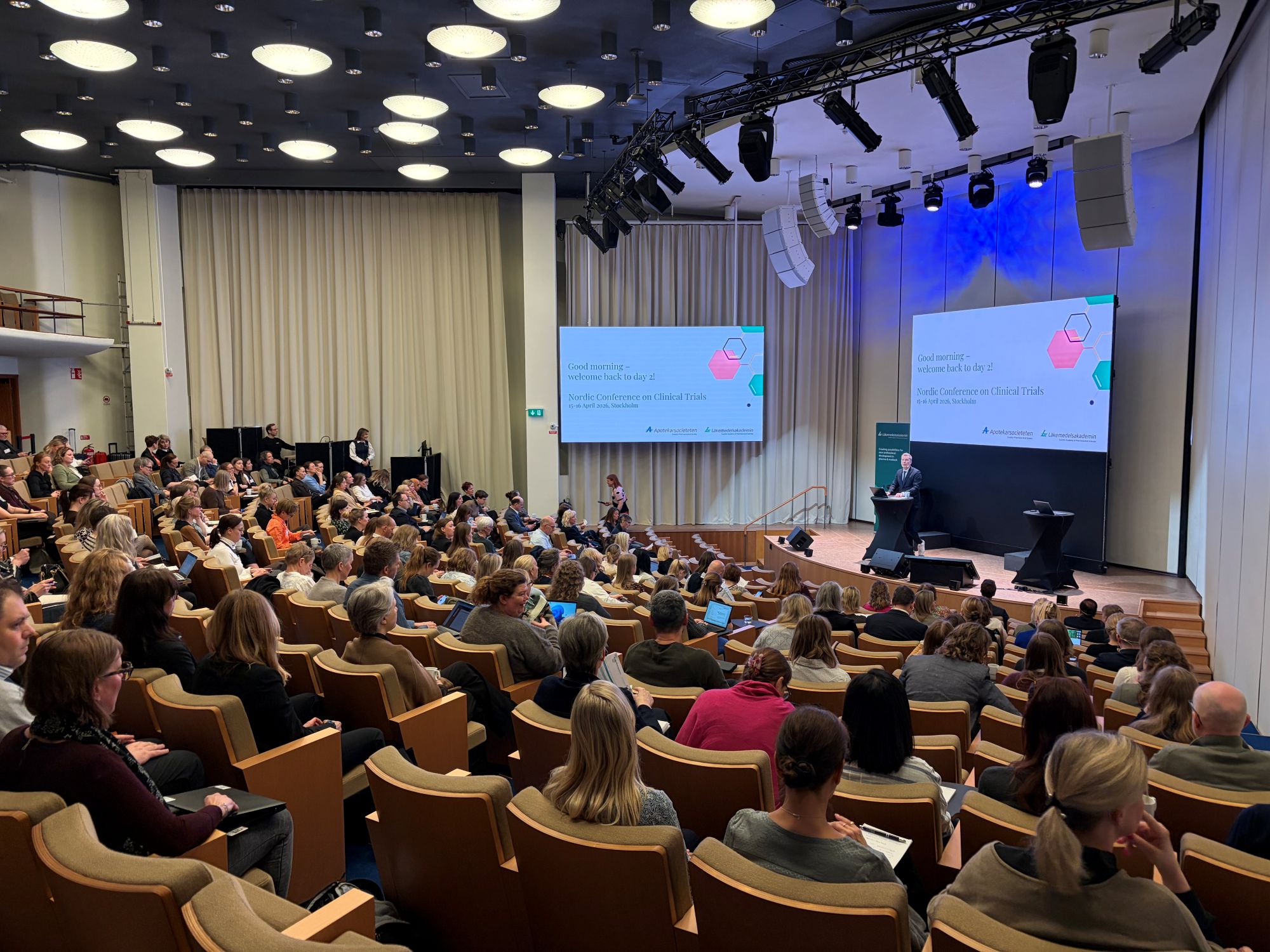 Bilde fra møtesalen: Publikum, konferansier, scene og skjerm